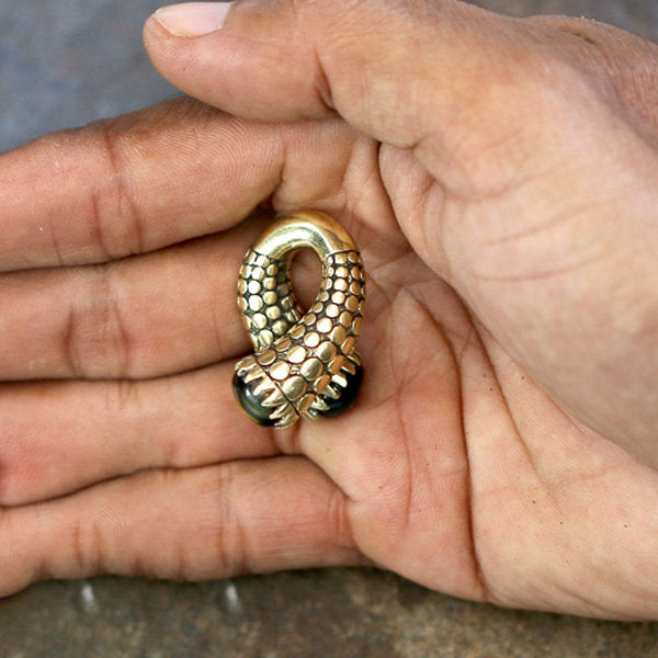 The photo features a hand holding the elegant TIMO Ear Weights in Gold with Tiger Eye from Kaya Movement. These metallic ear weights, designed to resemble a coiled snake with intricate scales, prominently showcase a tiger's eye stone. The ends of the snake design meet gracefully in the center. The background is softly blurred, drawing attention to the striking jewelry piece.